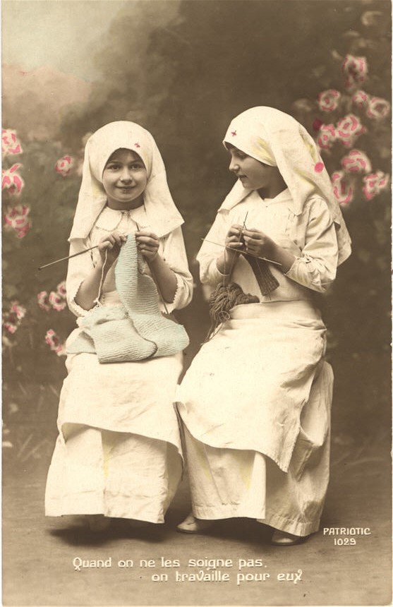 Two White girls dressed as nurses in white, sitting and knitting in a garden.