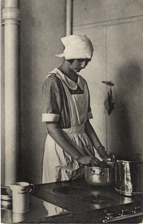 A White female in an apron and head covering at a stove, stirring a pot.