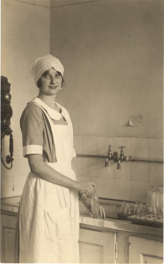 A White female (Princess Astrid)  in an apron and head covering washing bottles at the sink.