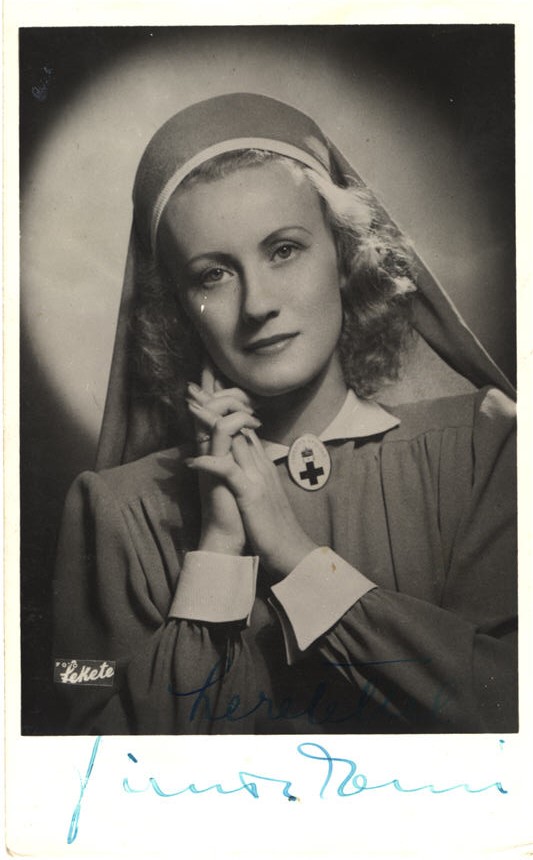 A White woman actress dressed nurse, hands clasped to the right of her face, looking at viewer.