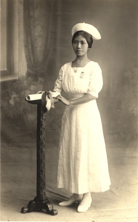 An Asian female nurse in white stands next to small podium with a diploma on it.
