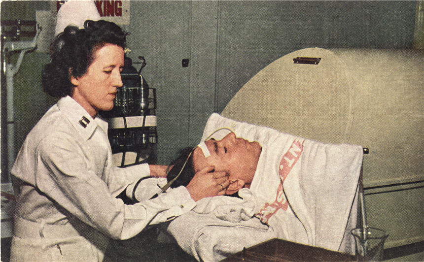 A White female nurse in white feels the temples of a White male patient in an iron lung.