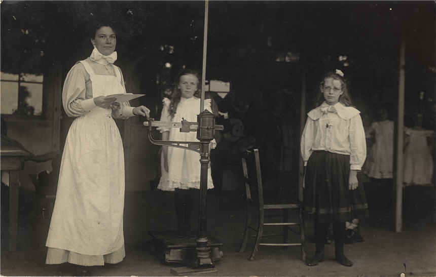 A White female nurse in white apron weighing a White girl as another White girl stands nearby.
