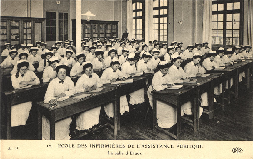 A large group of White female nurses in white sit in seven rows of desks in a classroom.