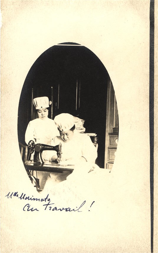 An Asian female nurse in white sits at work at a sewing machine, as another looks on.