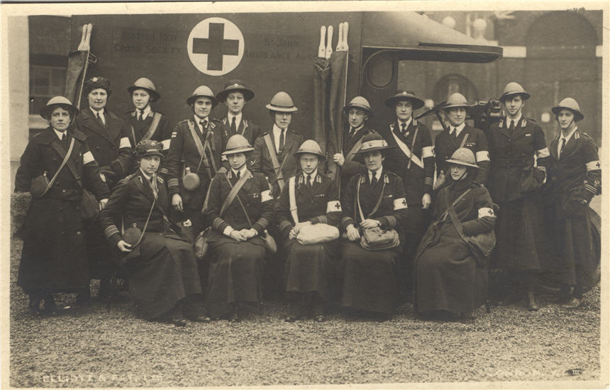 Sixteen White female nurses in dark military overcoats stand and sit in front of an ambulance.