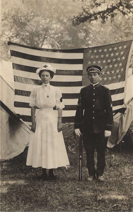 A White female nurse and a White male soldier in uniform stand in front of an American flag.