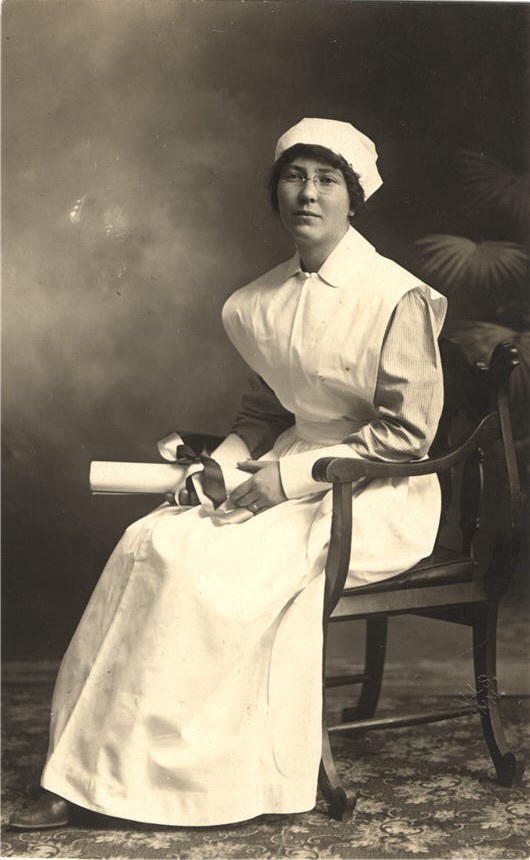 A White female nurse in white, sitting and holding a diploma.