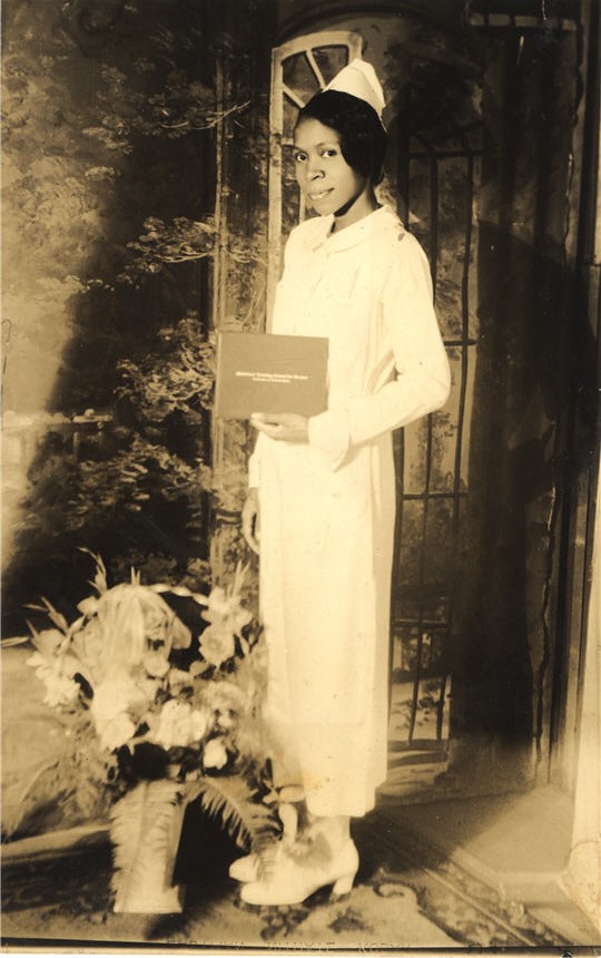 An African American female nurse in white holds diploma and looks at the viewer.