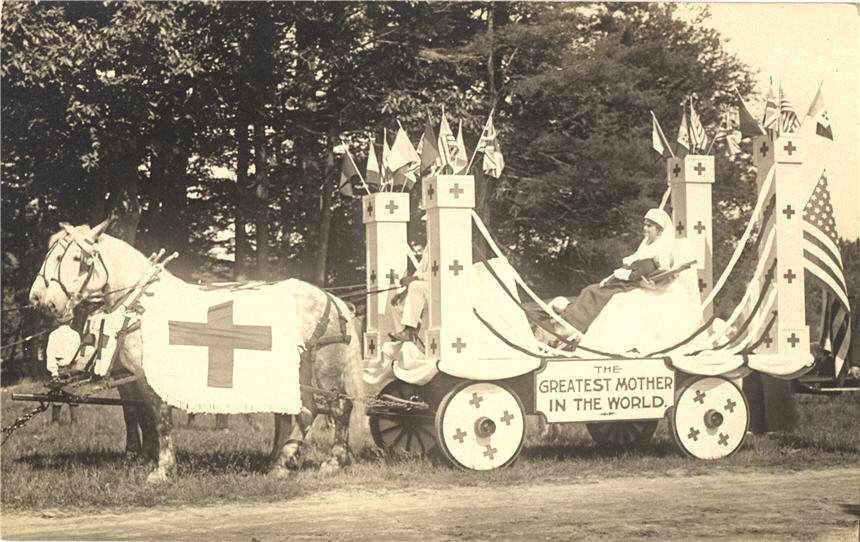 A White woman, dressed as a nurse, holds a baby in horse drawn wagon covered with Red Cross symbols.
