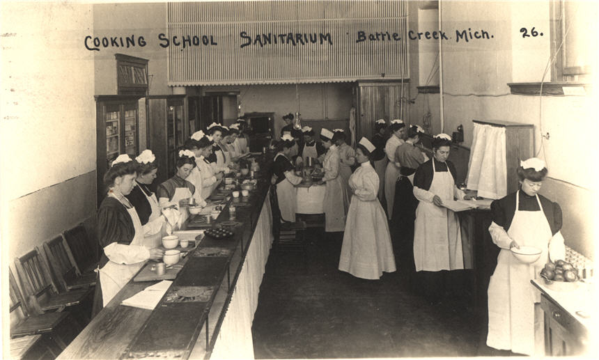 Twenty White female nurses in white aprons standing at various kitchen stations.