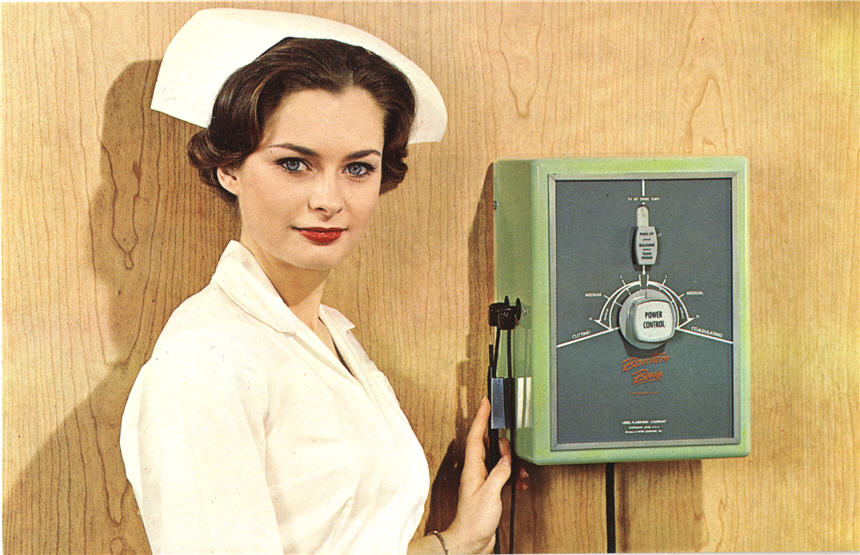 A White female nurse in white touches a medical device on a wall and looks at the viewer.