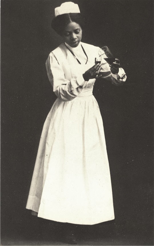 An African American nurse in white stands and pours liquid from one container to another.