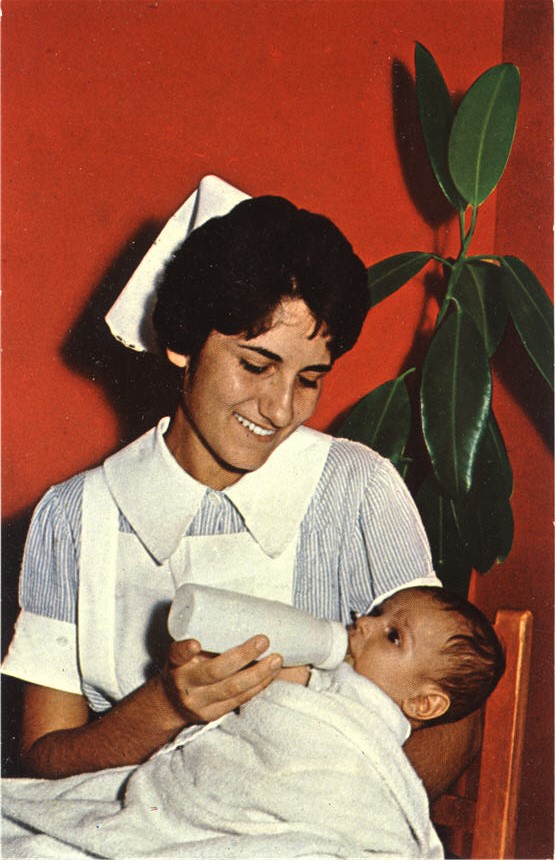 A White female nurse in white and blue sits and gives a White baby a bottle.