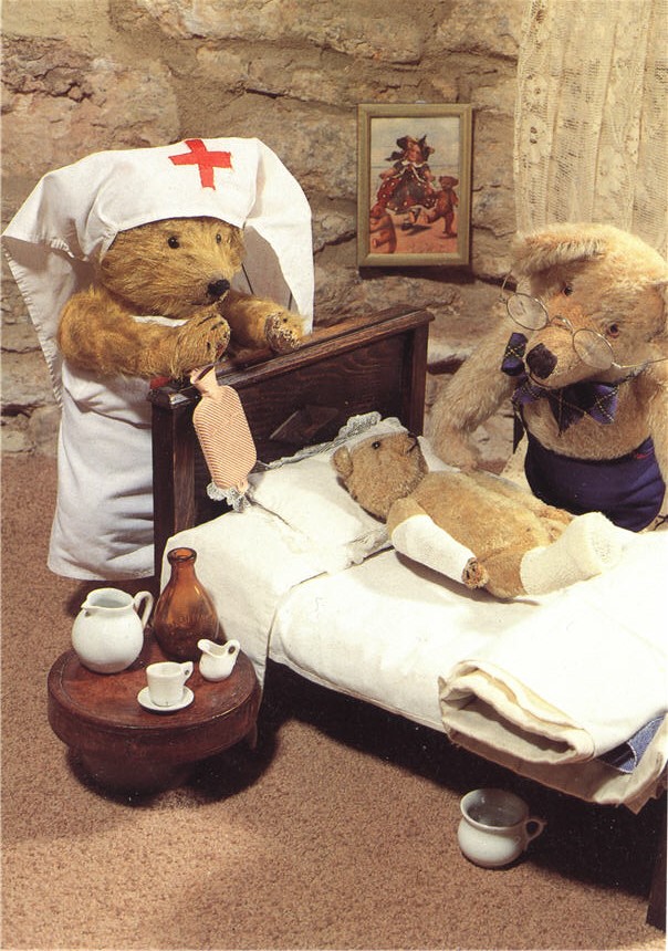 A teddy bear doll dressed as a nurse in white stands by a bear doctor as they examine a bear patient.