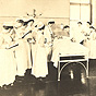 White male doctor leaning over a White patient. Seventeen White female nurses stand around the bed.