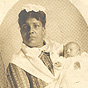 An African American female nurse in white holding a White infant, looking at the viewer.