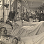 A WWI hospital ward. White male doctor stands to the left, White nurses and patients behind him.
