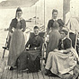 Four White women in brown and black, two are seated in chairs and the other two are standing.