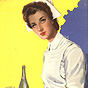 A White female nurse carrying a tray with a glass and bottle on it, looking at the viewer.