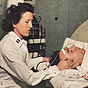 A White female nurse in white feels the temples of a White male patient in an iron lung.