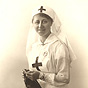 A White female nurse in white smiling, knitting, and looking at the viewer.