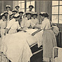 A group of White female nurses in white watch three others demonstrate bed making.
