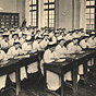 A large group of White female nurses in white sit in seven rows of desks in a classroom.