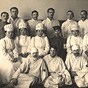 Six Asian female nurses in white, sit for group photograph with group of White male patients.