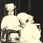 An Asian female nurse in white sits at work at a sewing machine, as another looks on.