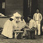 A group of White male patients in front of a building as a White female nurse nun tends to them.