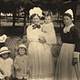 Three White female nurses in white and black with six White children, look at viewer.