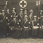 Sixteen White female nurses in dark military overcoats stand and sit in front of an ambulance.
