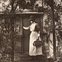 A White female nurse in white in front of a house with her hand on door, looking back at viewer