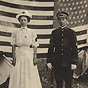 A White female nurse and a White male soldier in uniform stand in front of an American flag.