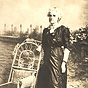 An elderly White female woman in black dress, standing next to a chair looking to the left.