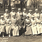 Nineteen White female nurses in white sit and stand for a group photo with a White male officer.