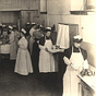 Twenty White female nurses in white aprons standing at various kitchen stations.
