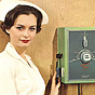 A White female nurse in white touches a medical device on a wall and looks at the viewer.