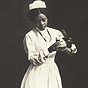 An African American nurse in white stands and pours liquid from one container to another.