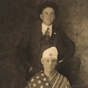 A White man wrapped in blanket and American flag sits in front of a stand White man in a suit.