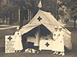 Three White female nurses stand outside a tent, as another woman tends to a man inside the tent.