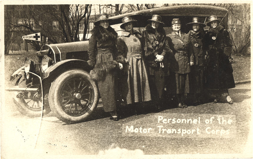 Six White female nurses standing in front of a car with a Red Cross symbol on a 'Motor Corps' flag.