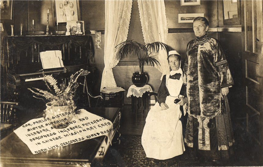 Two White missionary women inside a parlor. The seated woman on the left wears nursing clothes.