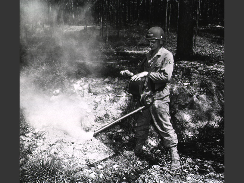 A photo of a white soldier in the woods spraying mist from an apparatus