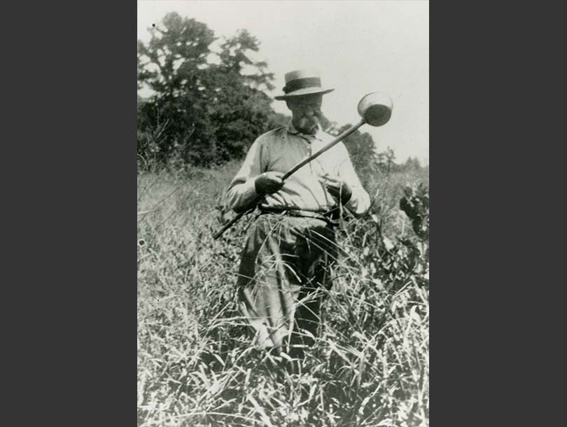 A photo of a white man surrounded by tall grass with a large scoop in hand