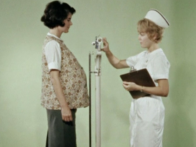 A pregnant white woman is weighed by a white female nurse