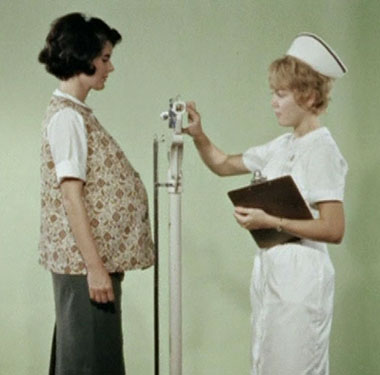 A pregnant white woman is weighed by a white female nurse