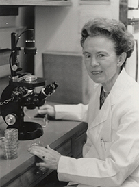Dr. Hattie Elizabeth Alexander, a White female in a lab coat next to a microscope and stack of petri dishes.
