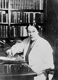 Dr. Virginia M. Alexander, a female in a lab coat writing at a desk with bookshelves behind her.